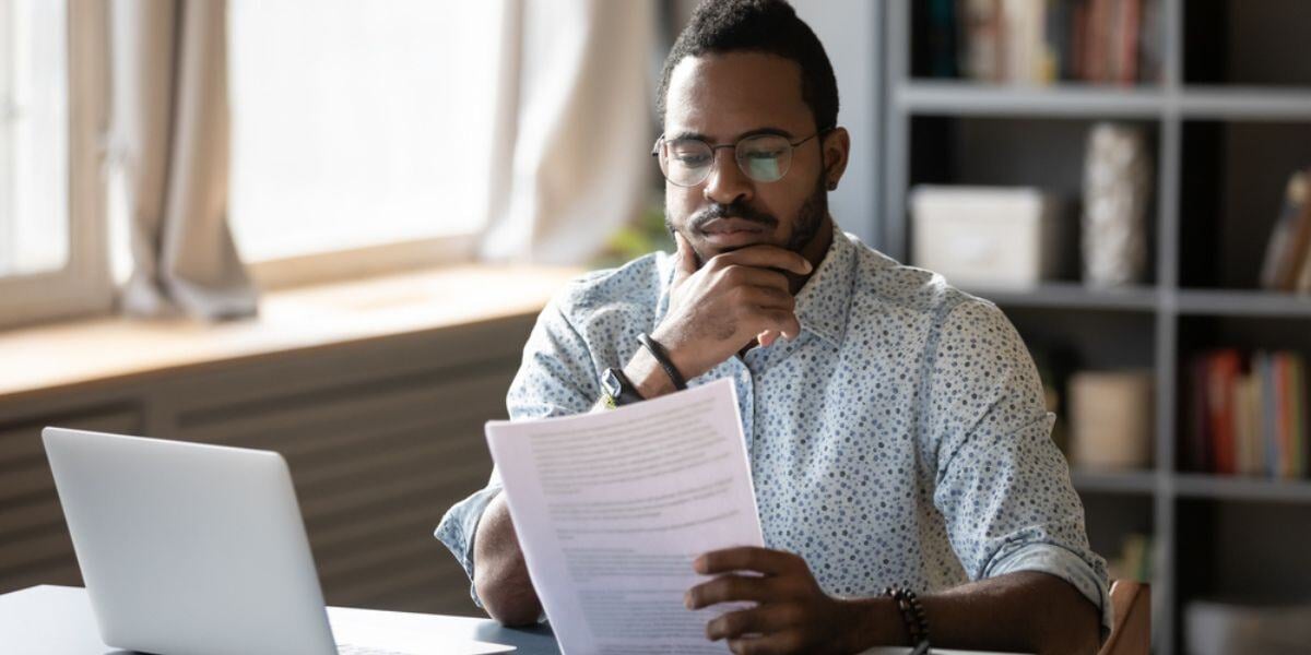 Pensive African American man sit at desk working on laptop reading paper document thinking or analyzing